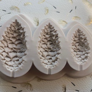 May include: A silicone mold for baking, featuring three detailed pine cone shapes. The mold is light gray and has a smooth, textured surface.