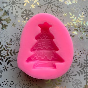 May include: Pink silicone mold for making a Christmas tree-shaped cake or other edible treats. The mold features a detailed design of a Christmas tree with a star on top.