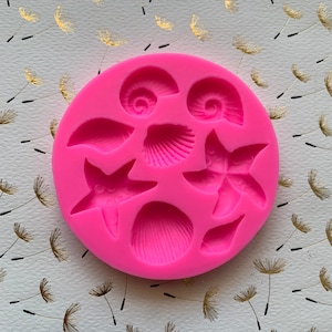 May include: Pink silicone mold with seven different shapes of seashells and starfish. The mold is designed for making edible decorations for cakes or other desserts.