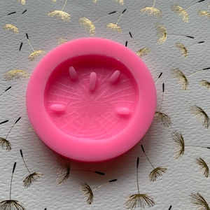 May include: Pink silicone mold with a raised design of a dandelion seed head. The mold is circular and has a smooth surface.