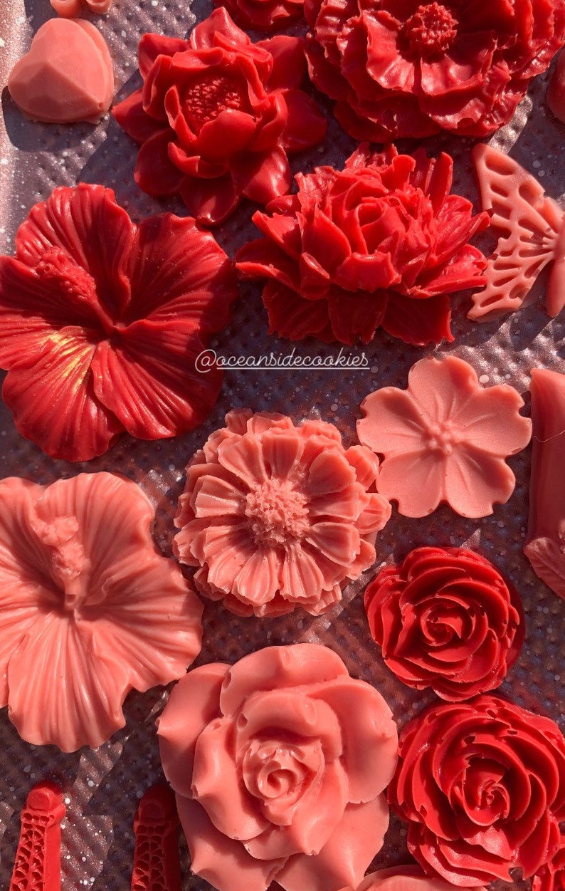 May include: Assortment of red and pink wax flower decorations. The flowers are arranged on a glittery surface. The flowers are in various shapes and sizes. The text "@oceansidecookies" is visible in the image.
