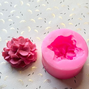 May include: Pink silicone mold for making a hydrangea flower. The mold is round and has a detailed design of a flower with multiple petals.