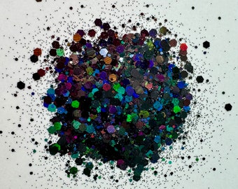 May include: A close-up of iridescent black, green, and blue glitter. The glitter is scattered on a white surface.