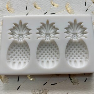 May include: A white silicone mold featuring three pineapple shapes. Each pineapple has a detailed crown and a textured body, perfect for creating decorative elements. The mold is designed for crafting and baking projects.