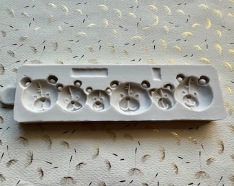 May include: A light gray silicone mold featuring five teddy bear faces of varying sizes. The mold has a rectangular shape with a small handle on the left side. The bear faces have detailed features.