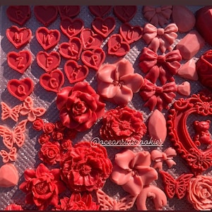 May include: A collection of red and pink wax melts in various shapes, including hearts, flowers, butterflies, and a picture frame with a teddy bear inside. The wax melts are arranged on a glittery surface. The text "@oceansidecookies" is visible in the lower right corner of the image.