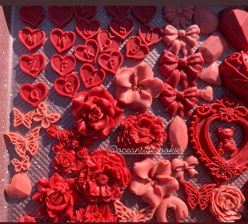 May include: A collection of red and pink wax melts in various shapes, including hearts, flowers, butterflies, and a picture frame with a teddy bear inside. The wax melts are arranged on a glittery surface. The text "@oceansidecookies" is visible in the lower right corner of the image.
