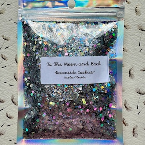 May include: A clear plastic bag filled with silver, black, and iridescent glitter. The bag has a label that reads "To The Moon and Back - Oceanside Cookies - Naples, Florida."