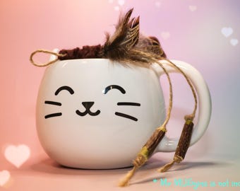 May include: White ceramic mug with a cat face design and a brown feather and twine accent. The mug has a handle and is on a pink and purple background.