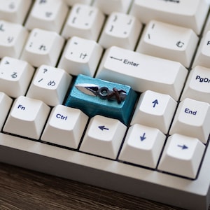 Pode incluir: Teclado mecânico branco com uma tecla azul e prateada com um pequeno design de espada. O teclado tem uma caixa branca e teclas brancas.