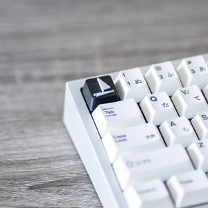 May include: A white mechanical keyboard with Japanese characters and a black key with a sailboat icon. The keyboard has a gray frame and is set on a wooden surface. The keys are white with blue and black lettering.