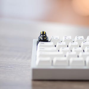 May include: A white mechanical keyboard with a small, black and gold figurine on one of the keys. The keyboard sits on a light gray wooden surface, with a blurred background.