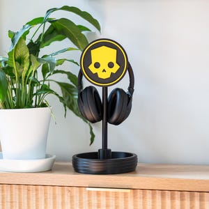 Puede incluir: Auriculares negros sobre la oreja en un soporte negro. El soporte tiene un logotipo de calavera amarilla sobre un fondo negro. El soporte está sobre una superficie de madera, con una planta en maceta en el fondo.