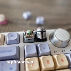 May include: A white mechanical keyboard with custom keycaps featuring a Grogu figure and a Mandalorian helmet. The keyboard also has a silver volume knob and the word "alphalocked" in the lower left corner.