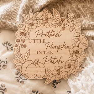 May include: A wooden sign with the text "Prettiest Little Pumpkin in the Patch", framed by a floral wreath and a pumpkin illustration. The sign is made of light-coloured wood and has a scalloped edge.