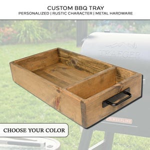 Op de afbeelding: Een op maat gemaakte BBQ-lade van lichtbruin hout met een verdeeld interieur. Het heeft zwarte metalen hardware en de tekst "CUSTOM BBQ TRAY" en "CHOOSE YOUR COLOR". De lade is ontworpen voor buiten grillen.
