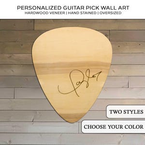 May include: Oversized guitar pick wall art made of hardwood veneer. The light-colored wood grain is visible. The pick features a signature and the text "TWO STYLES" and "CHOOSE YOUR COLOR".