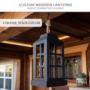 May include: A black wooden lantern with a rectangular frame and a grid pattern. It hangs from a rope and hook. The text "CUSTOM WOODEN LANTERNS" and "CHOOSE YOUR COLOR" are displayed on the image.