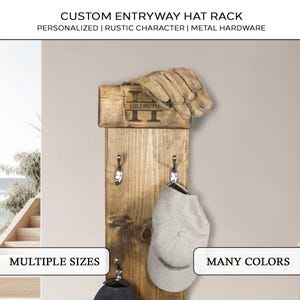 May include: A rustic wooden entryway hat rack with metal hooks. The rack is personalized with the name "HILDRETH" and the letter "H". A pair of gloves and two hats hang from the hooks. The text "CUSTOM ENTRYWAY HAT RACK" is at the top.