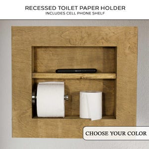May include: A recessed wooden toilet paper holder with a cell phone shelf. The holder is made of light brown wood and has a rectangular frame. It features a shelf for a cell phone and space for two rolls of toilet paper. The text "RECESSED TOILET PAPER HOLDER INCLUDES CELL PHONE SHELF" is at the top.