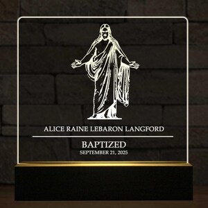 May include: A rectangular LED light with a black base and a white border. The light features a white illustration of a religious figure with outstretched arms. The text "ALICE RAINE LEBARON LANGFORD" and "BAPTIZED SEPTEMBER 21, 2025" are also displayed.