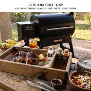 Può includere: Un vassoio rettangolare in legno per barbecue con una sezione divisa e manici in metallo nero. Il vassoio contiene un pollo arrosto, rosmarino fresco e tre barattoli di spezie in vetro etichettati Sea Salt, Pepper e Smokey Mesquite Rub.