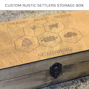May include: Custom rustic Settlers storage box with engraved game board elements. The wooden box features a dark stain on the sides and a lighter wood top with engraved game icons and text. The box has a black metal clasp.