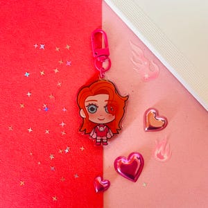 May include: A pink and red cartoon character keychain with a red heart, a pink heart, and a pink flame design. The keychain is made of clear acrylic and has a shiny finish.
