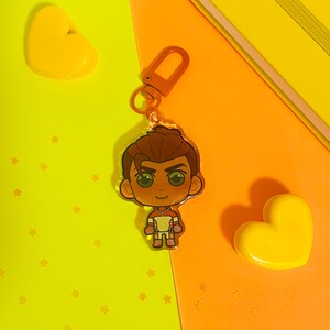 May include: A brown and orange cartoon character keychain with a white and red racing suit. The character has brown hair and green eyes. The keychain is on a yellow and orange background.