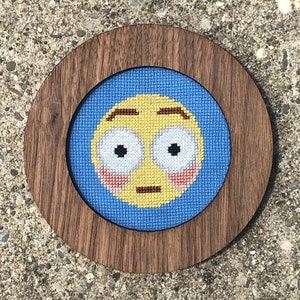 May include: A wooden framed embroidery hoop with a blue background and a yellow face with wide eyes and blushing cheeks. The face is embroidered with a simple design.