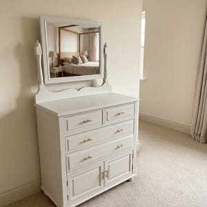 May include: A light grey wooden chest of drawers featuring a rectangular mirror attached to the top. The unit has two small top drawers, two full-width middle drawers, and two lower cabinet doors, all accented with small, metallic gold-toned handles.