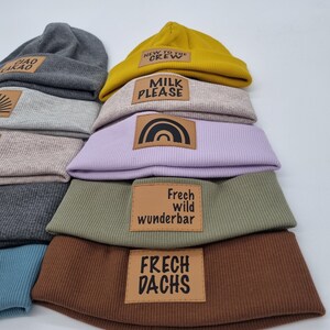 Könnte beinhalten: Ein Stapel von acht Strickmützen in verschiedenen Farben, jede mit einem anderen braunen Lederflicken mit einem einzigartigen Spruch. Die Sprüche lauten "New to the Crew", "Milk Please", ein Regenbogen, "Frech wild wunderbar" und "Frech Dachs".