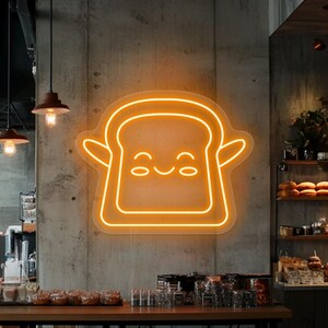 May include: A neon sign in the shape of a smiling piece of toast with arms, glowing orange against a concrete wall. The sign is in a cafe setting, with wooden tables and stools visible in the foreground.