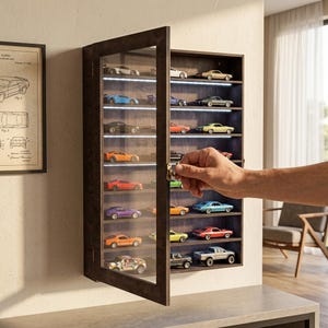May include: A wall-mounted dark wood display case with a glass door and internal LED lighting, featuring multiple shelves filled with various miniature toy cars in assorted colors. The case has a brass latch and a hinged door that opens outward.