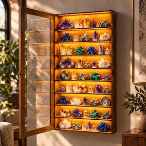 May include: A rectangular wooden wall-mounted display cabinet with a glass door, featuring eight illuminated shelves filled with various colourful mineral specimens, including amethyst clusters, quartz points, and metallic pyrite formations.
