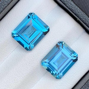 May include: Two vibrant blue topaz gemstones, cut in an emerald shape, are displayed in a white velvet box. The gemstones are faceted and reflect light, showcasing their brilliance.