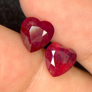 May include: Two heart-shaped, faceted, deep red gemstones. The gems are held in a hand, with a dark background. The facets catch the light, creating a sparkling effect. These gemstones could be used for jewellery.