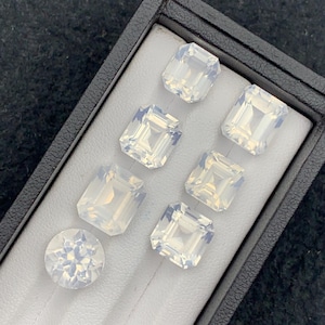 Könnte beinhalten: Eine Sammlung opalisierender Edelsteine in verschiedenen Formen, darunter Smaragd- und Rundschliffe, präsentiert in einer schwarzen Box mit grauem Innenraum. Die Edelsteine haben eine milchig-weiße Farbe und reflektieren das Licht.