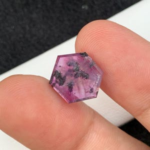 May include: A faceted, hexagonal gemstone with a deep pink hue and dark inclusions. The gem is held between two fingers, showcasing its polished surfaces and unique internal patterns. The gemstone is a natural mineral.