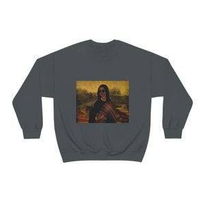 May include: A dark grey sweatshirt with a print of a woman wearing sunglasses and a shawl, standing in a landscape setting. The print is reminiscent of a famous painting.