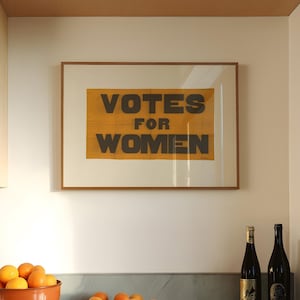 gs print shoppe, gsprintshoppe.com, vintage-style suffrage poster art print featuring bold black text 'VOTES FOR WOMEN' on an orange-yellow banner, framed in minimalist wood and displayed in bright modern interiors