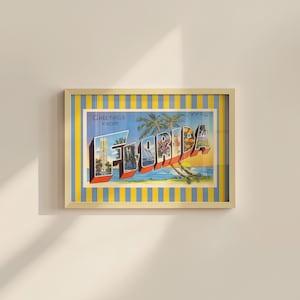 K&ouml;nnte beinhalten: Gerahmte Vintage-Postkartenkunst mit den Worten "Greetings from Florida" in Rot, Wei&szlig; und Blau. Die Postkarte zeigt Illustrationen von Palmen, einem Segelboot und einem Sonnenuntergang. Der Rahmen hat eine goldene Oberfl&auml;che und einen blau-gelb gestreiften Rand.