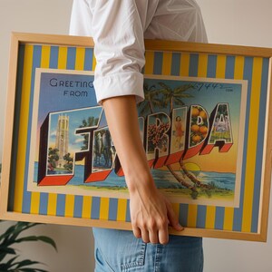 K&ouml;nnte beinhalten: Gerahmtes Vintage-Postkartenkunstwerk mit der Aufschrift "Greeting from Florida". Das Kunstwerk zeigt ein farbenfrohes Design mit dem Wort "FLORIDA" in gro&szlig;en, stilisierten Buchstaben, das ikonische Florida-Bilder wie Palmen, Orangen und einen Sonnenuntergang zeigt. Der Rahmen ist hellholzfarben.
