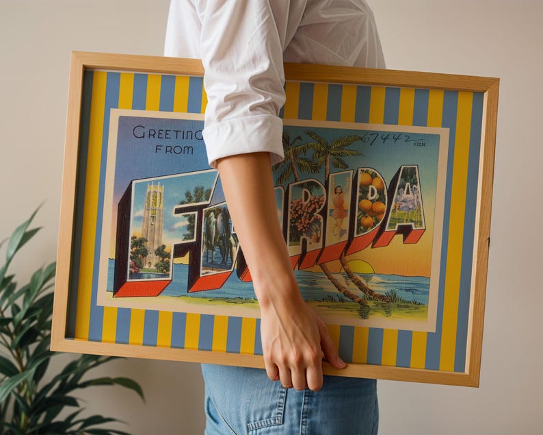 K&ouml;nnte beinhalten: Gerahmtes Vintage-Postkartenkunstwerk mit der Aufschrift "Greeting from Florida". Das Kunstwerk zeigt ein farbenfrohes Design mit dem Wort "FLORIDA" in gro&szlig;en, stilisierten Buchstaben, das ikonische Florida-Bilder wie Palmen, Orangen und einen Sonnenuntergang zeigt. Der Rahmen ist hellholzfarben.
