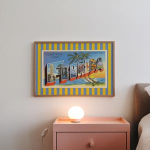 K&ouml;nnte beinhalten: Gerahmte Vintage-Postkartenkunst mit dem Wort "FLORIDA" in gro&szlig;en, farbenfrohen Buchstaben. Das Kunstwerk zeigt einen blauen Himmel, Palmen und einen Sonnenuntergang. Der Rahmen hat einen gelb-blauen Streifen.