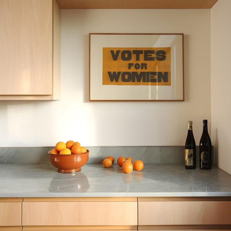 gs print shoppe, gsprintshoppe.com, vintage-style suffrage poster art print featuring bold black text 'VOTES FOR WOMEN' on an orange-yellow banner, framed in minimalist wood and displayed in bright modern interiors