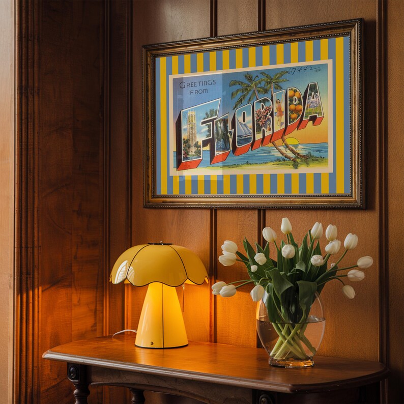 K&ouml;nnte beinhalten: Ein gerahmter Vintage-Postkarten-Druck mit den Worten "Greetings from Florida" im Retro-Stil. Eine gelbe Lampe mit einem gewellten Schirm steht auf einem Holztisch neben einer Vase mit wei&szlig;en Tulpen.