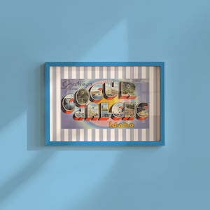 Puede incluir: Una postal vintage enmarcada con un marco azul. La postal dice "Greetings from Coeur d'Alene Idaho" en una fuente retro. El fondo de la postal es un patrón de rayas. El marco está colgado en una pared azul claro.