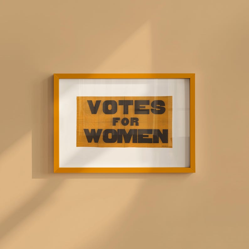gs print shoppe, gsprintshoppe.com, vintage-style suffrage poster art print featuring bold black text 'VOTES FOR WOMEN' on an orange-yellow banner, framed in minimalist wood and displayed in bright modern interiors