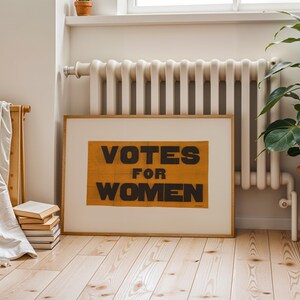 gs print shoppe, gsprintshoppe.com, vintage-style suffrage poster art print featuring bold black text 'VOTES FOR WOMEN' on an orange-yellow banner, framed in minimalist wood and displayed in bright modern interiors